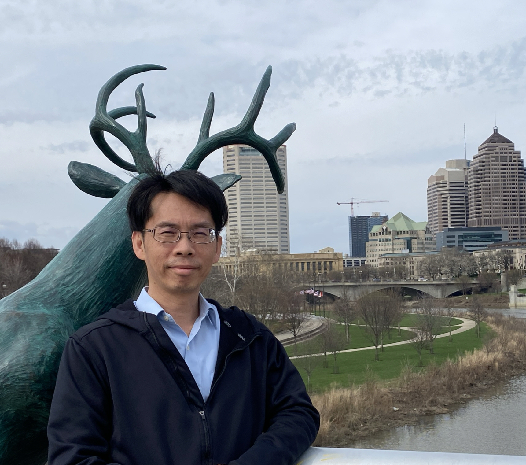 An Asian man with glasses wearing a sky blue collared shirt and black sweater stands in front of a statue of an antelope with a city skyline in the background