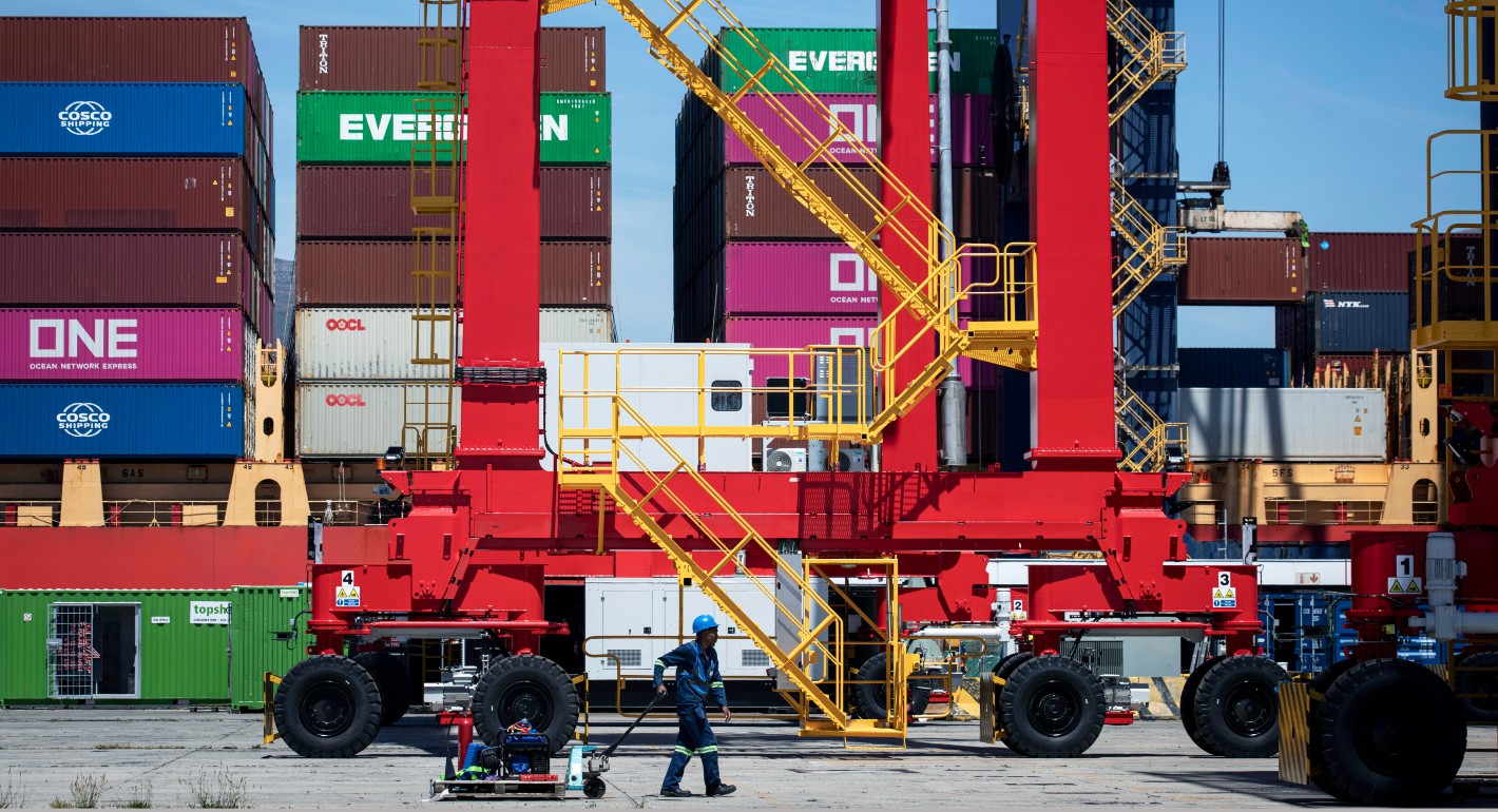 A Black man pulls a trolley. He is small in the bottom center of the frame; in the background are stacks of large, colorful shipping containers and the parts of a large crane or similar piece of equipment.