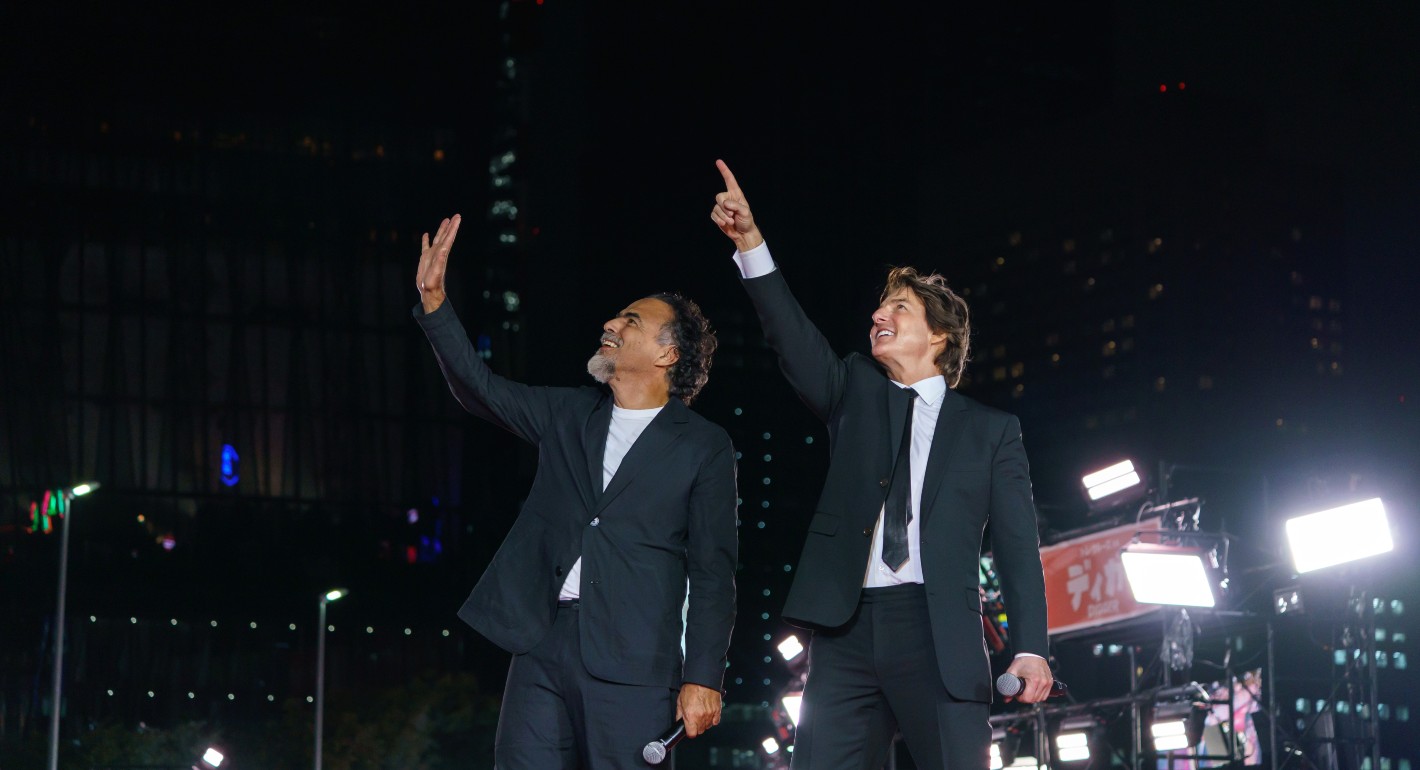 Two men wave and point to the sky against a dark background