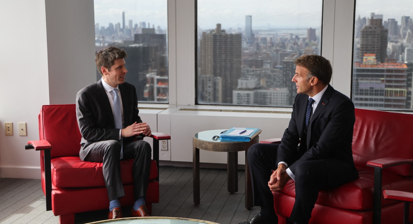 Two men sit on red chairs in front of a window with a NYC city skyline.