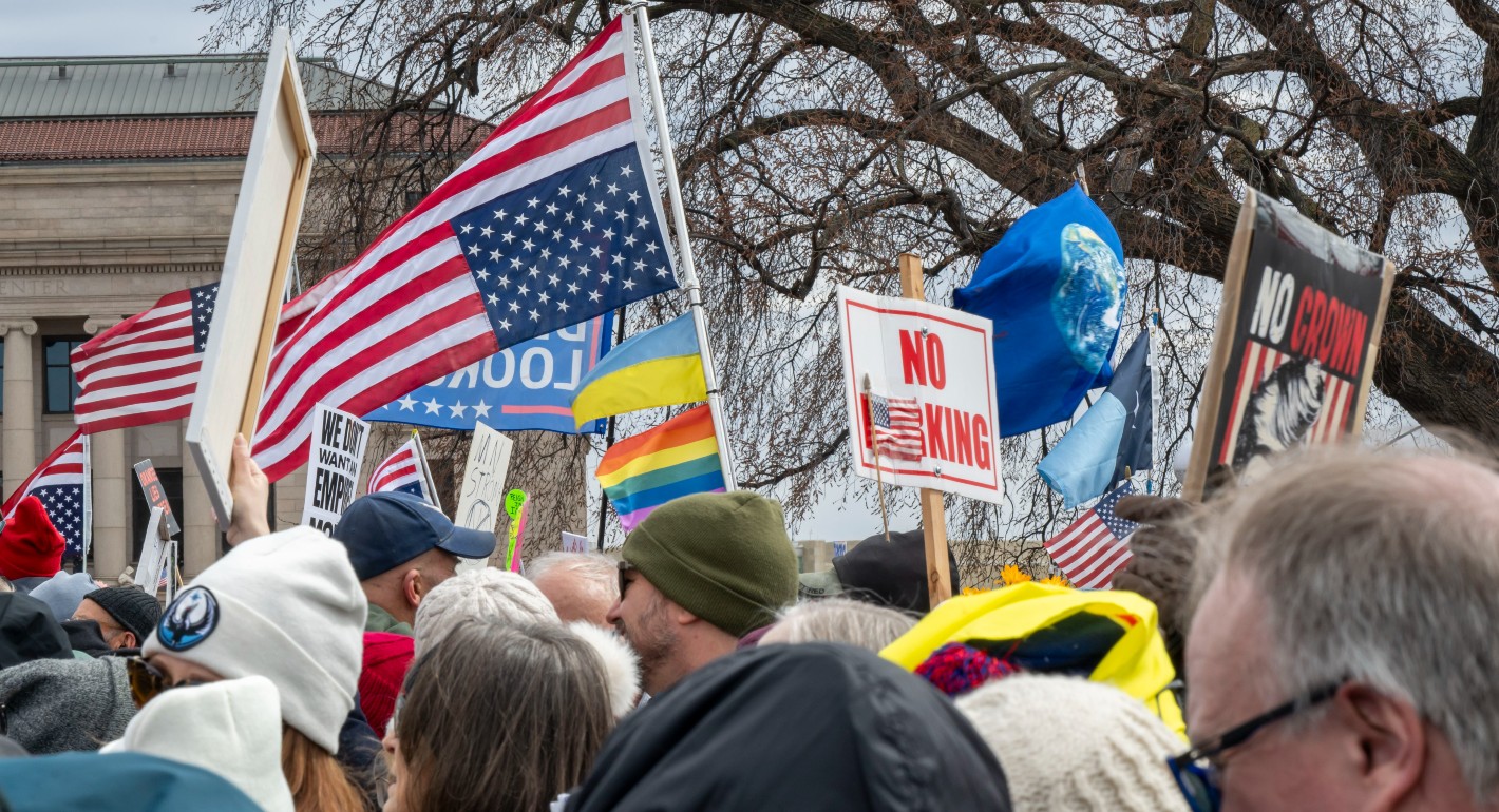 The tops of people's heads. Raised above their heads are "No Kings" signs, an upside-down American flag, and a rainbow flag.
