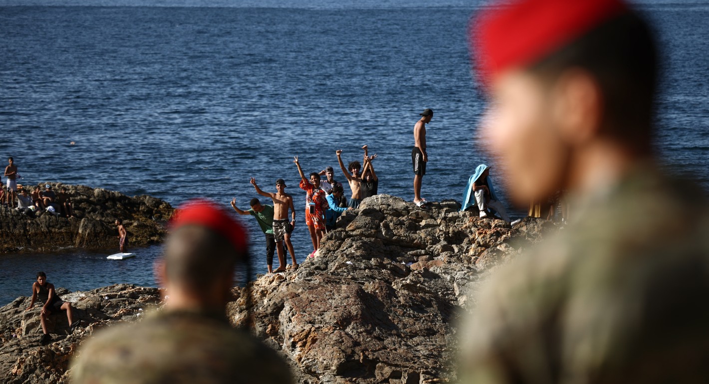 People standing on rocks in the ocean near the shore, with soldiers in the foreground