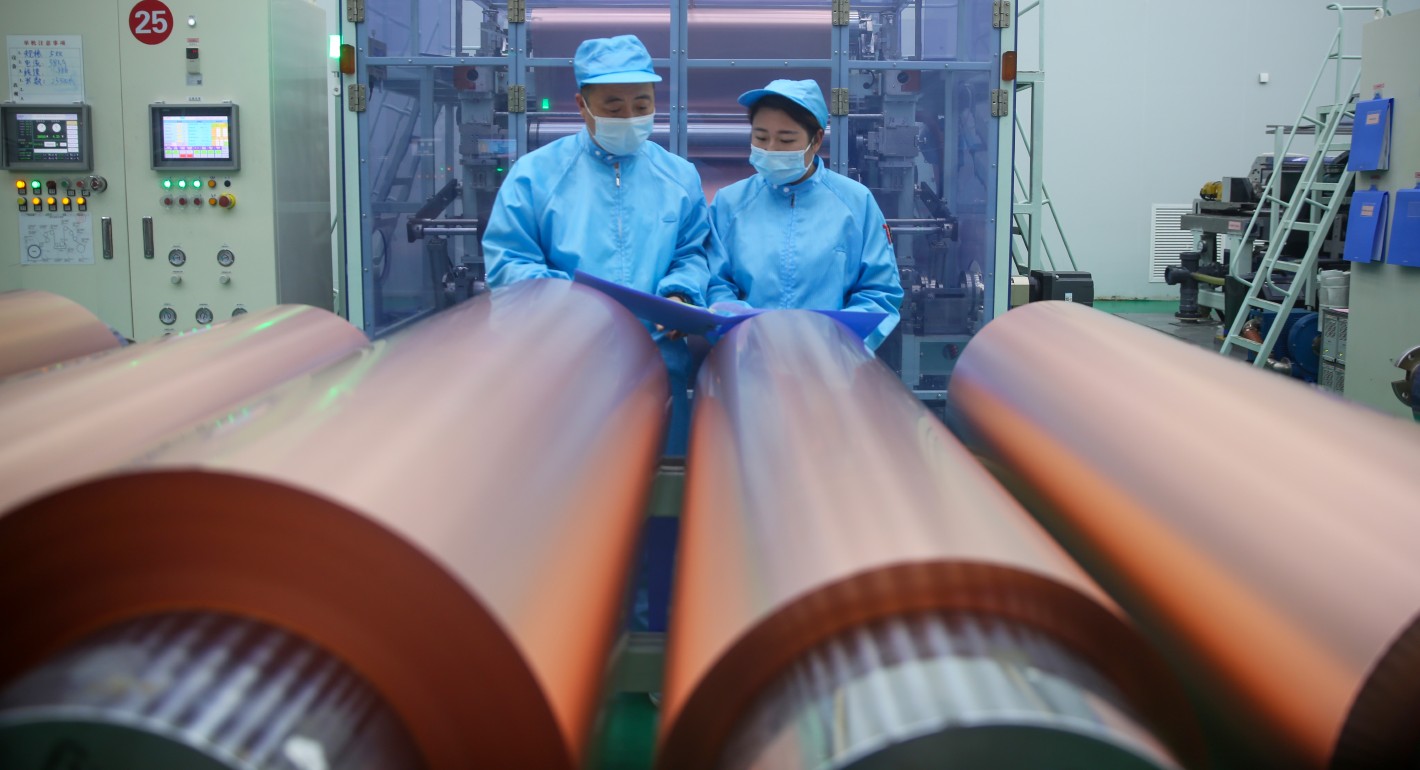 Two people in blue PPE standing over large metal rolls