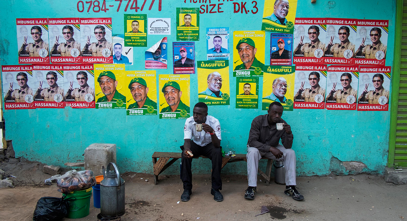 Men take a tea break in front of a wall with election posters near a polling station in Dar es Salaam on October 25, 2015, during the Tanzanian presidential election.