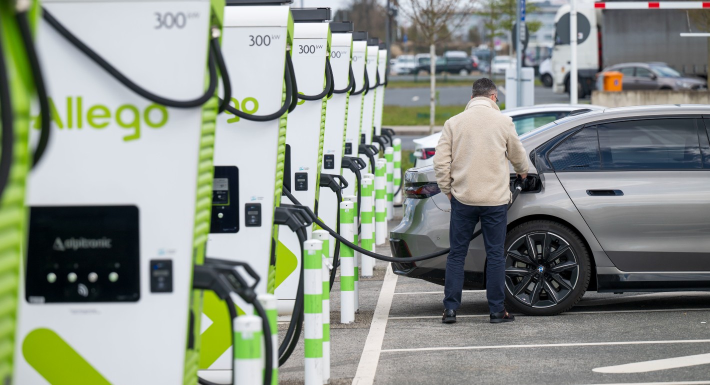 A White man in a tan jacket stands with his back to the camera, plugging in an electric car to a row of green and white chargers.