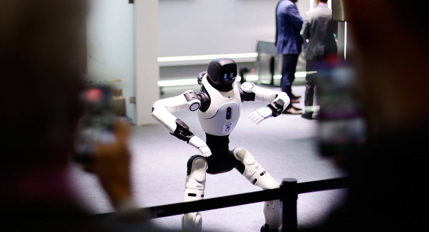 A white humanoid robot with a black head and hips stands behind a rope barrier. It stands with its feet apart, knees bent, and elbows raised at its sides.