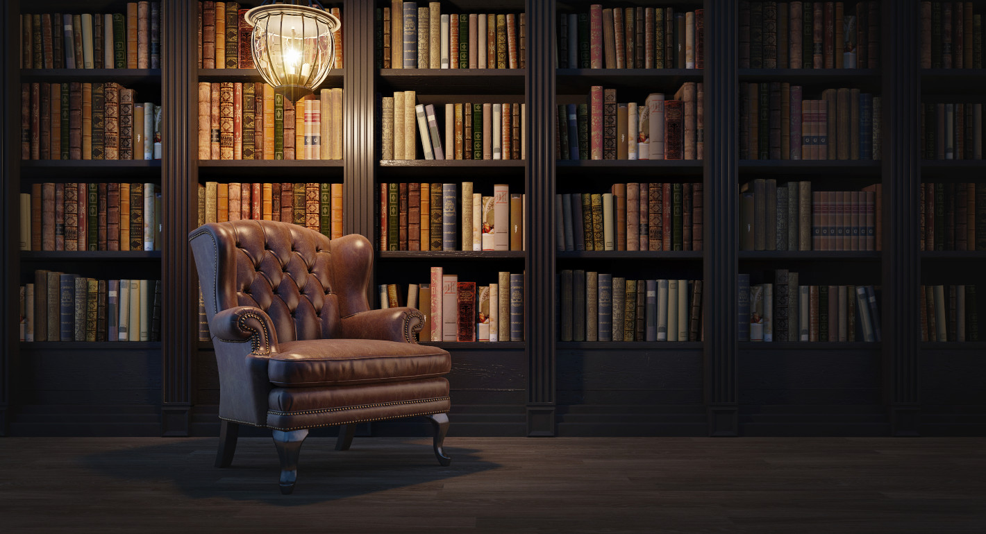 Photo of a leather armchair below a hanging lamp in front of a dark wood bookshelf.