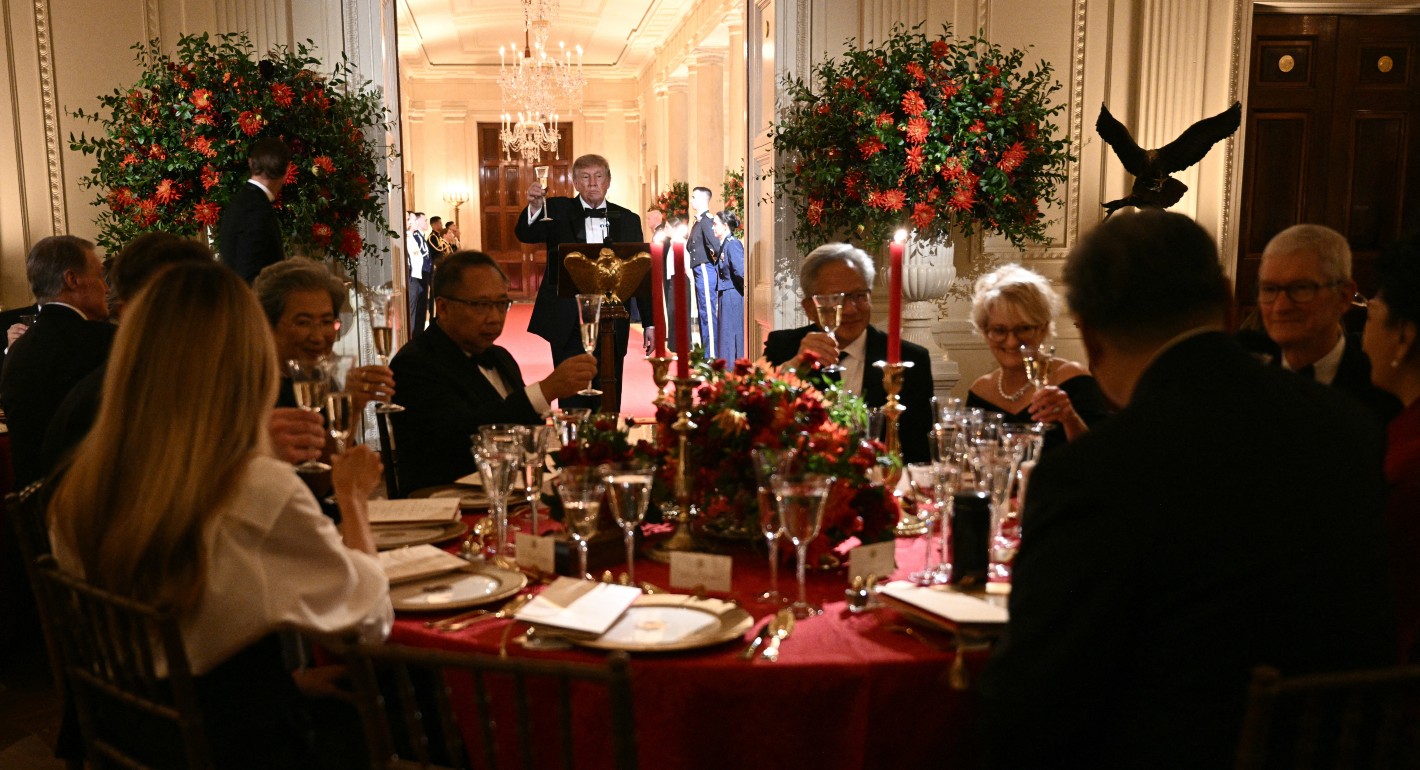 Trump holding a glass of champagne up in a toast, with a table of guests in the foreground