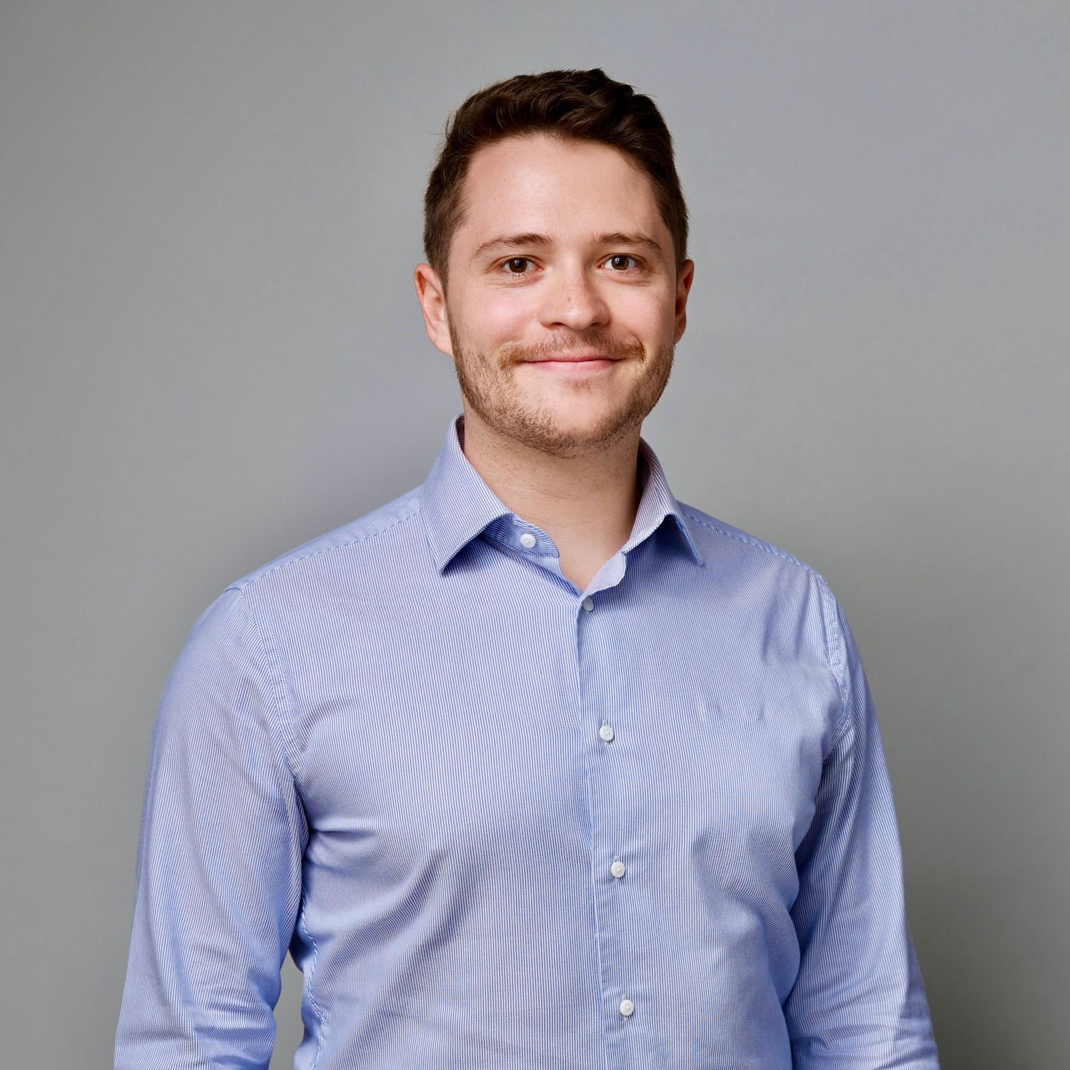 A white man with brown-red hair wears a blue collared shirt and smiles without teeth at the camera