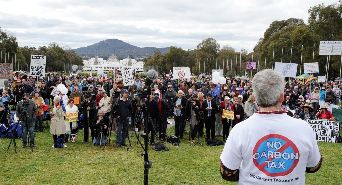 A man stands with his back to the camera in front of a large crowd holding protest signs standing on the lawn in front of Australia's Parliament. The back of the man's shirt reads "No Carbon Tax" with a red X.