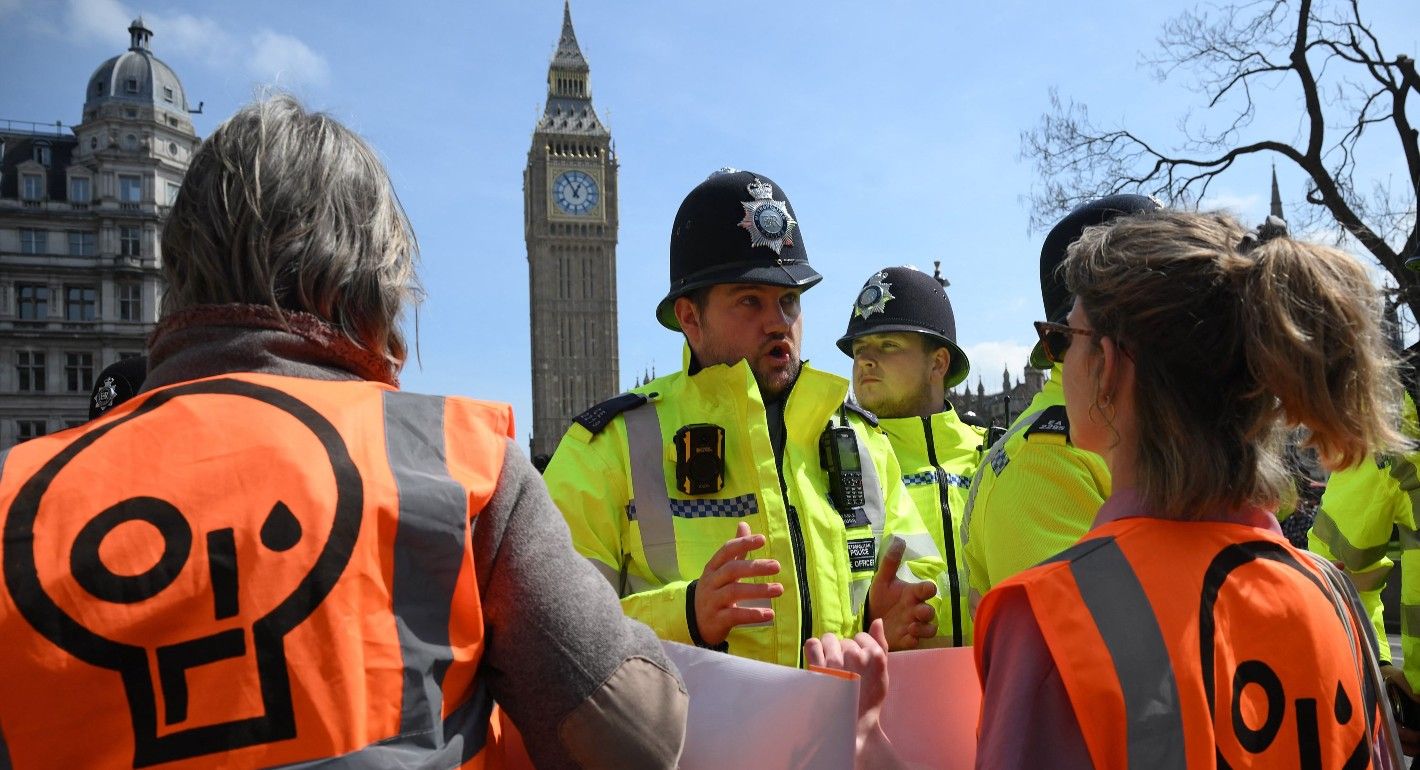 Police in bright yellow jackets stand face to face with protesters in bright orange. The police face the camera and the protesters' backs are to the camera
