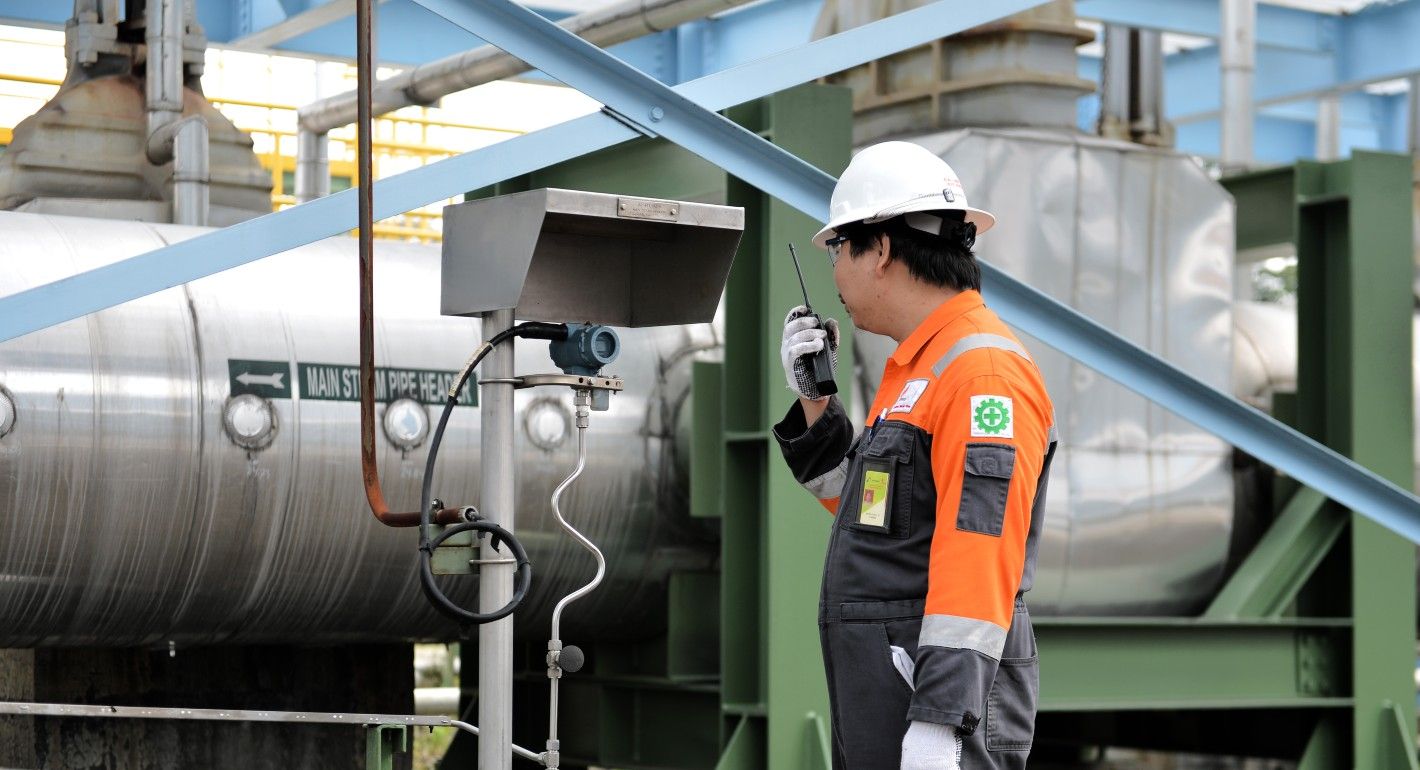 A man in a white construction hat and orange construction uniform holds a walkie talkie. He stands in front of large geothermal machinery and looks at a meter of some kind.