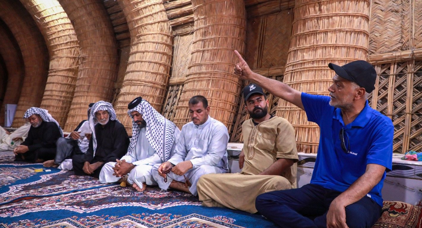 Several Iraqi men in traditional attire sit cross legged on a rug.