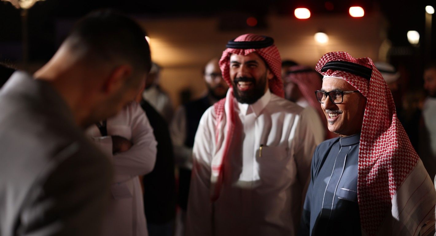 Two young men in ghutras talk to a third man whose back faces the frame. They talk and laugh under bright lights in a dark room.