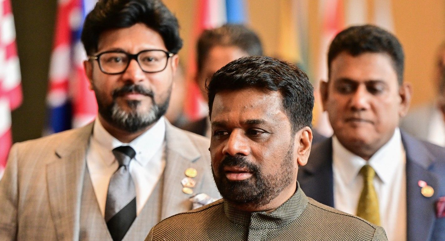 Close up of the face of Sri Lankan president Dissanayake as he walks. Two men are standing behind him, and there is a row of flags in the distant background.