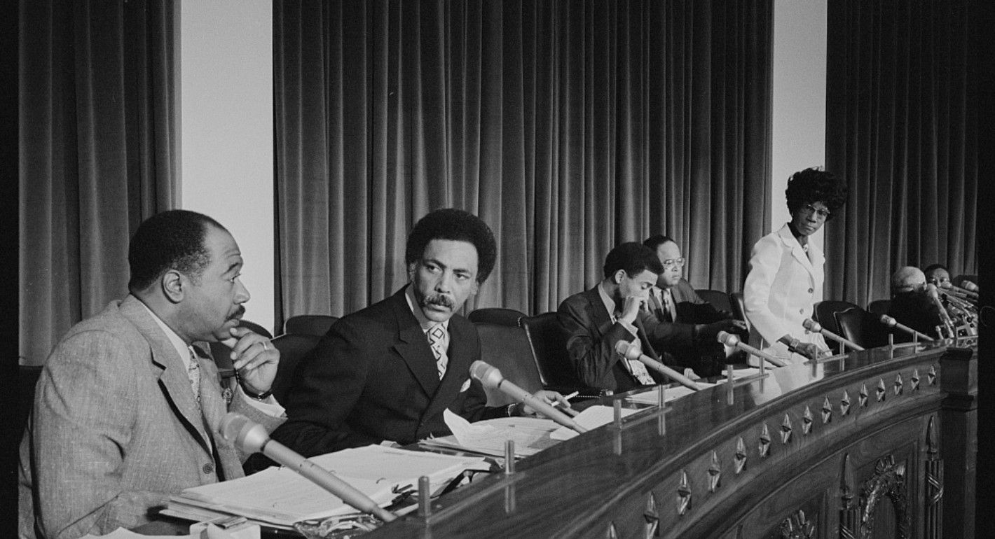 Photo of members of the Congressional Black Caucus sitting at a desk covered in papers.