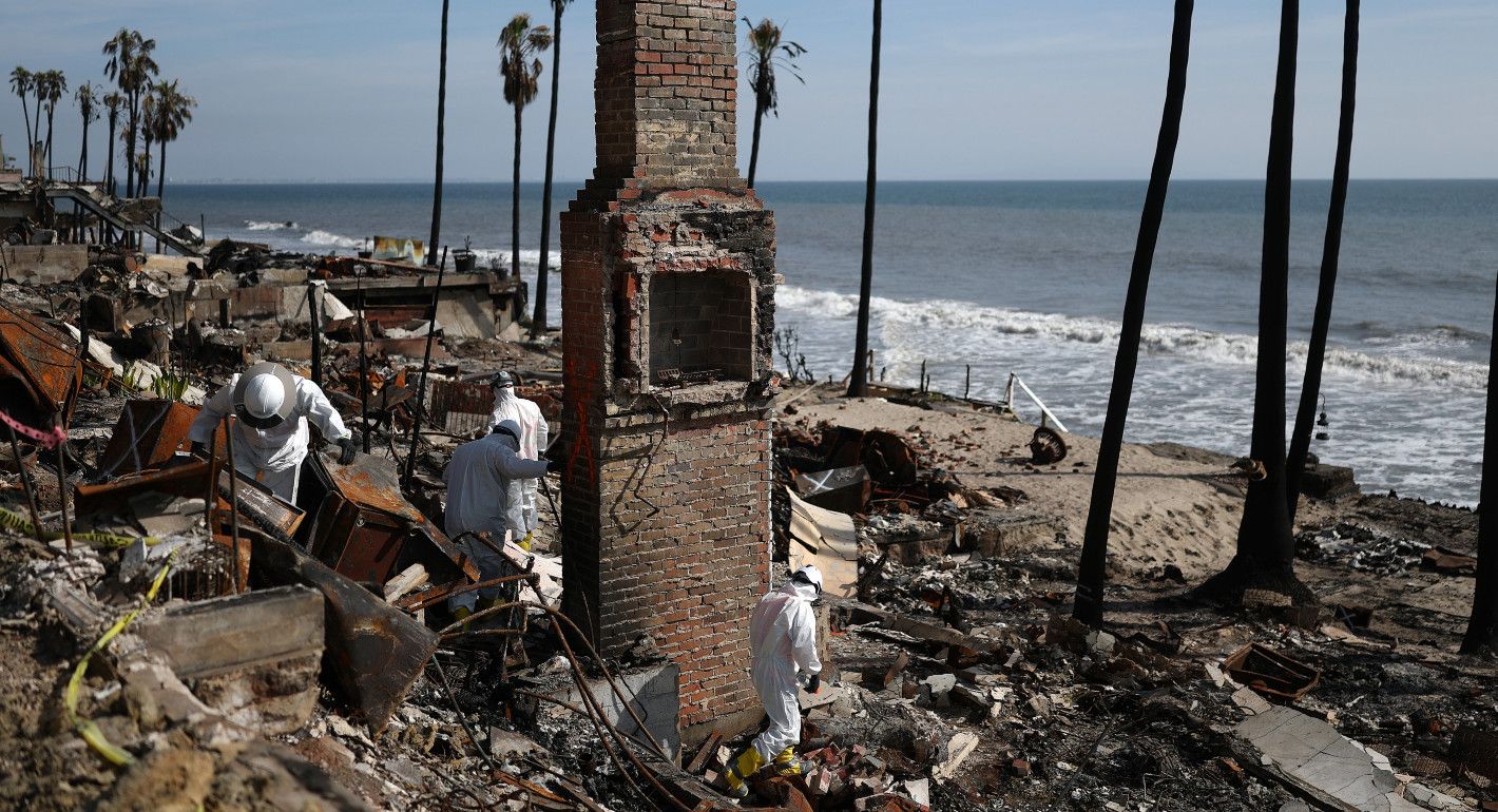 People in hazmat suits combing through a burned down house with only a chimney remaining