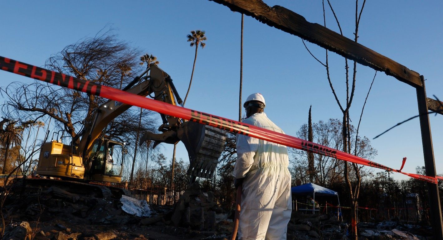Person in a hazmat suit directing a bulldozer to clean up debris from a burned-down home