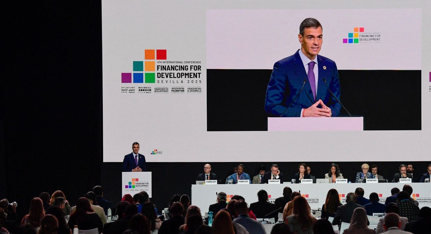 A man in a suit stands behind a podium. There is a screen behind him with the FFD4 logo and a video projection of himself. In the foreground are rows of people sitting facing him listening to the speech.