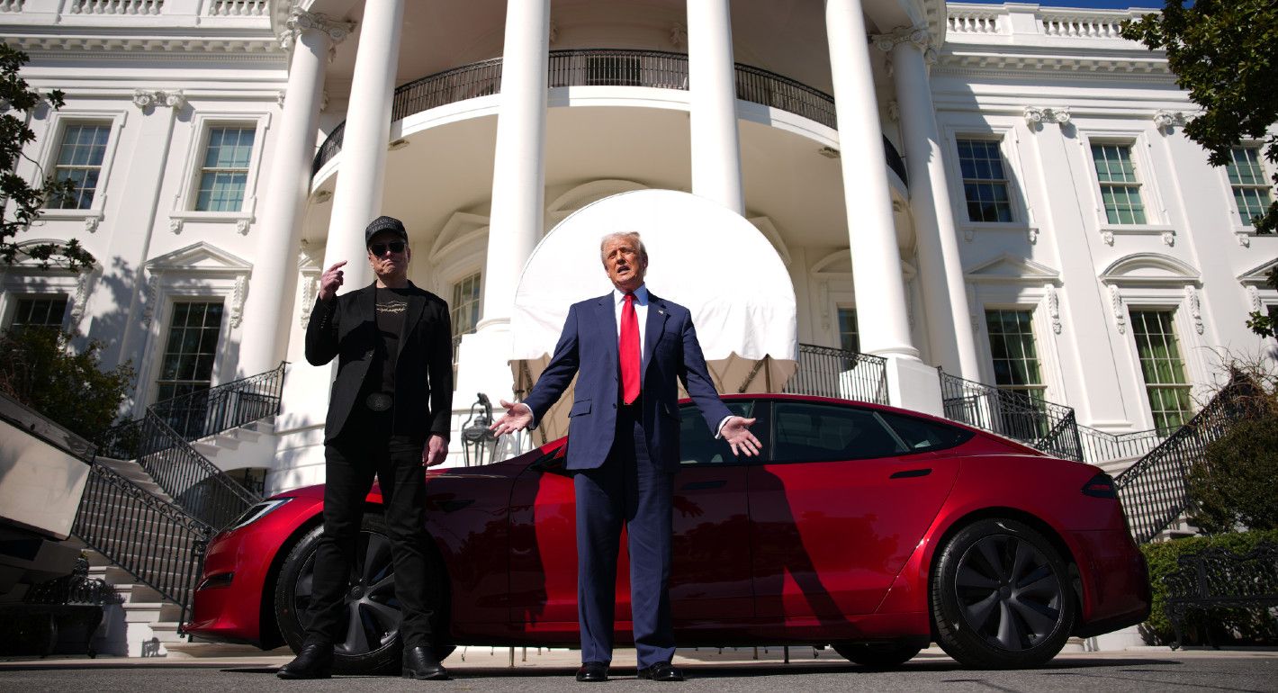 Photo of Donald Trump and Elon Musk standing in front of the White House, facing the camera, with a red Tesla behind them.