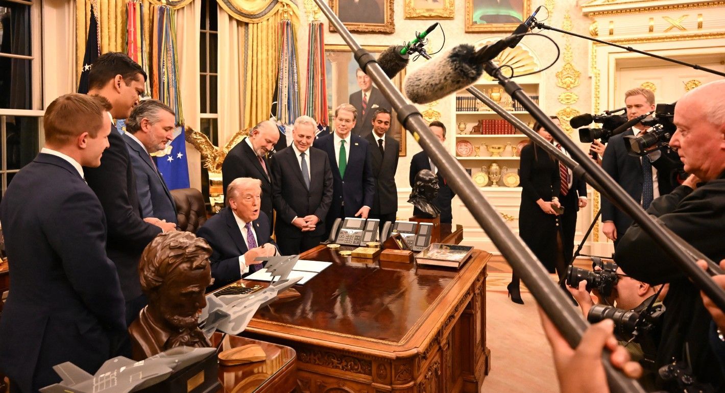 Trump sitting at a desk, surrounded by men in suits on one side and journalists on the other