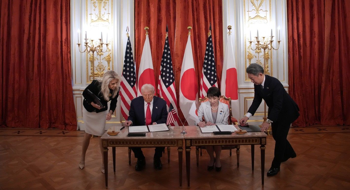 two leaders sitting at a table signing documents, with flags in the background and aides at their sides
